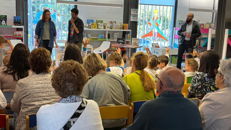 Audience of children and adults listening to two presenters on a small stage in a bright library or bookstore, with colorful shelves of books behind them.