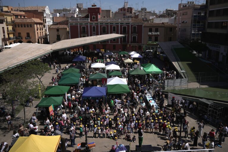 Top-down view of a crowded outdoor market in a plaza with green and white tents, people browsing stalls and a small stage on the side.