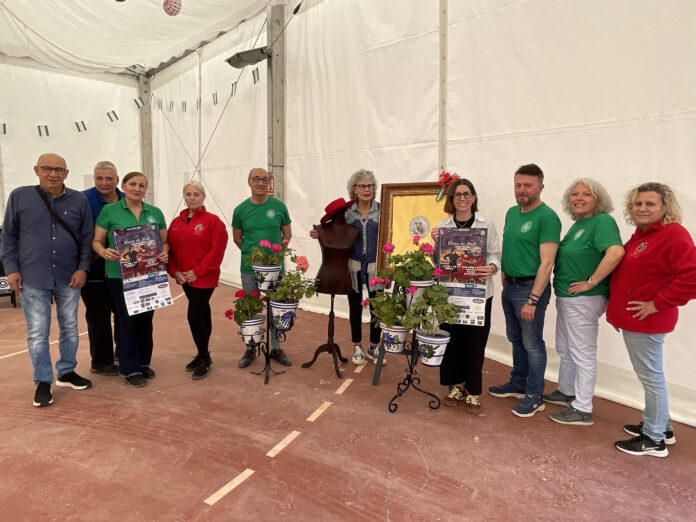 Group of ten people in a white tent posing for a photo; several wear green shirts, others red, holding posters and potted flowers.
