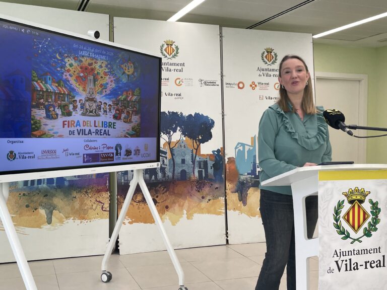 Woman in a teal blouse speaks at a white podium with a microphone, in front of a large colorful 'Fira del Llibre de Vila-real' poster screen and city crest backdrop.
